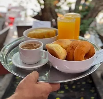Bandeja de inox servida com café, suco de laranja, uma tigela de salgados dourados e outra menor ao fundo.