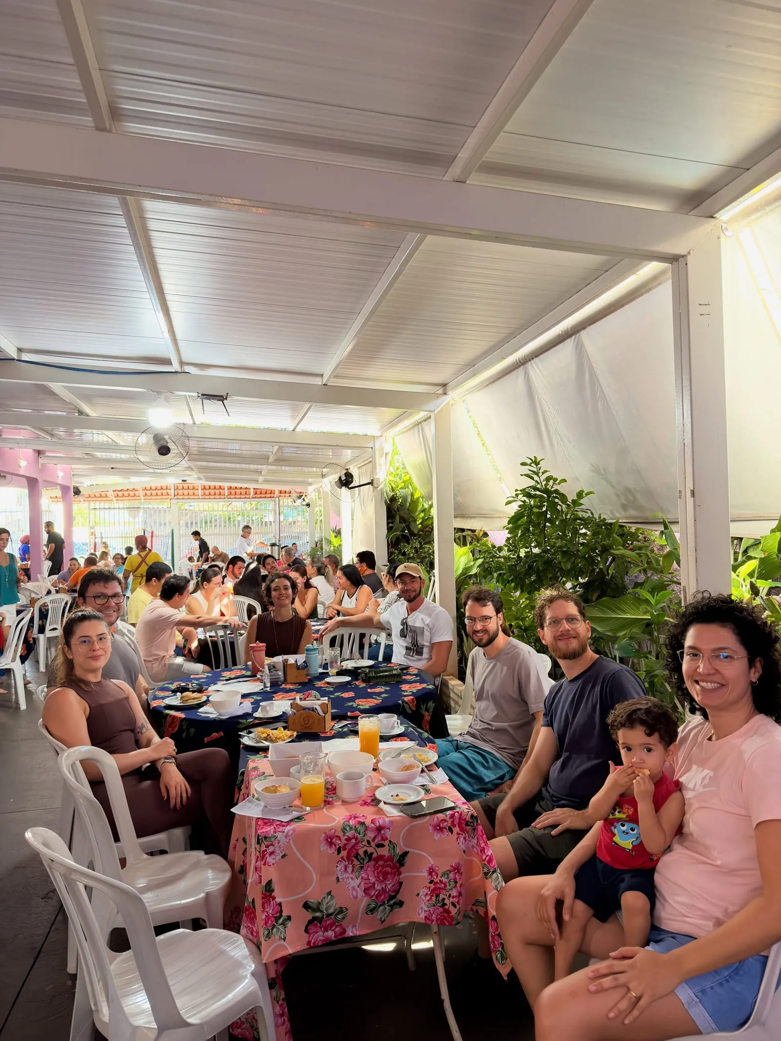 Área coberta da casa lotada num domingo de manhã: famílias em mesas redondas com toalhas floridas.