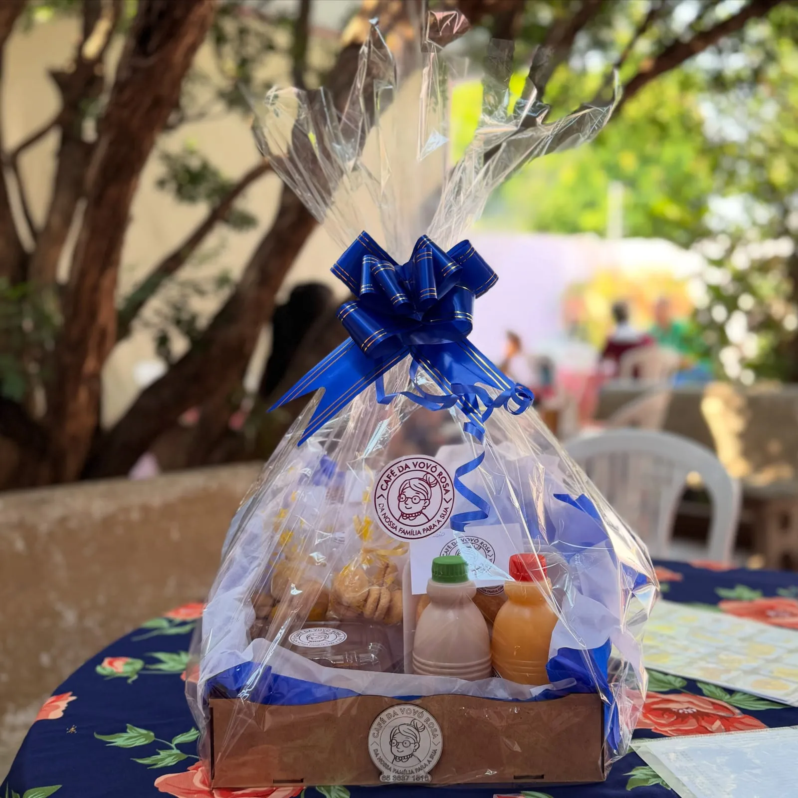 Cesta de café da manhã embalada em celofane com laço azul, com biscoitos, garrafinhas de suco e o selo do Café da Vovó Rosa.