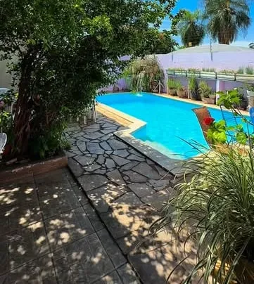 Quintal da casa: caminho de pedra entre árvores, piscina e vasos de plantas junto ao muro lilás.