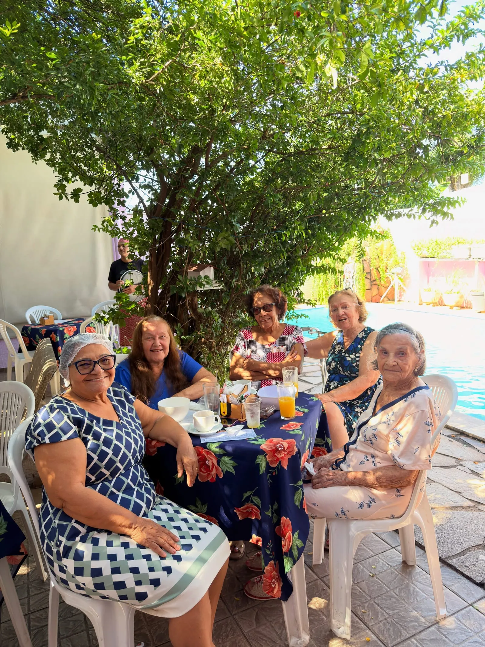 Grupo de amigas tomando café numa mesa com toalha azul florida, sob uma árvore, ao lado da piscina.