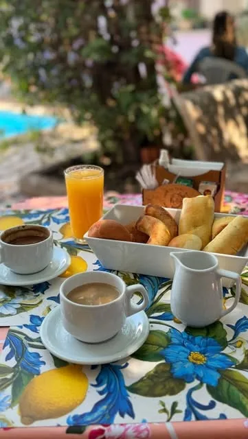 Mesa com toalha de chita azul e limões: duas xícaras de café com leite, leiteira, suco de laranja e uma travessa de salgados, com o jardim e a piscina ao fundo.