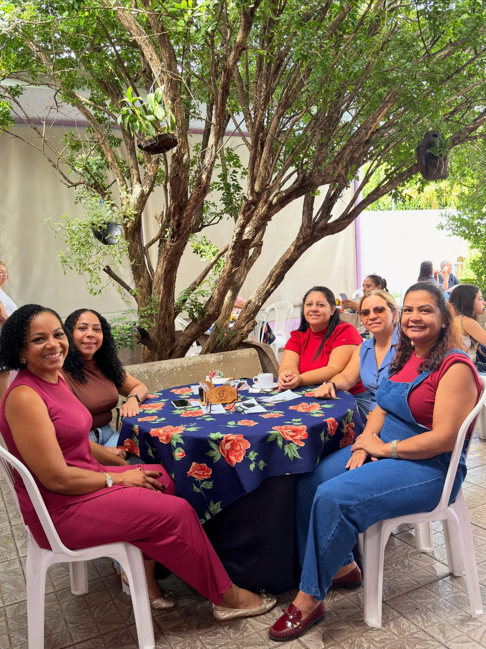 Mesa cheia de amigas sorrindo à sombra de uma árvore, com toalha azul de flores vermelhas.